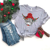 Shaggy Cow Hat Screen Print Heat Transfer, graphic t-shirt with a shaggy cow wearing a red and green striped Christmas hat with pom-pom, placed on a flat surface along with distressed jeans, a small holiday gift box, and Christmas decorations.