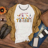 Give Thanks Gnomes Sublimation Transfer T-Shirt, white t-shirt featuring colorful gnome illustrations and the words 'give thanks'  design on the front, laid on a wooden surface with casual accessories.