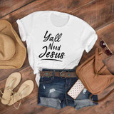Y'all Need Jesus Sublimation Transfer, white T-shirt featuring the text 'Y'all Need Jesus' in black lettering, accompanied by casual summer accessories like a straw hat, sunglasses, distressed denim shorts, a brown belt, a leather handbag, and sandals, laid out on a wooden surface.