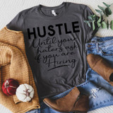 Hustle til the Haters Black Screen Print Heat Transfer, casual outfit with a graphic t-shirt, jeans, boots, a knit sweater, and decorative pumpkins, flat lay style.