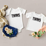 Twins Double Or Nothing Sublimation Transfer, white baby bodysuits with black text, one with long sleeve and one with short sleeve, displayed with blue shorts, flowers, and fabric accessories.