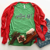 Gettin' Lit Screen Print Heat Transfer, festive holiday-themed green t-shirt with 'gettin' lit!' text and string of colorful Christmas lights, paired with a red cardigan, denim shorts, and holiday accessories.