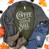 Coffee Warms The Soul Heat Transfer T-shirt, dark green with white text, surrounded by a gray knit cardigan, blue ripped jeans, beige ankle boots, and autumn leaves and pumpkins.