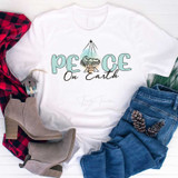 Peace On Earth Sublimation Transfer, white T-shirt with a Christmas-themed design of a nativity scene and the text 'Peace On Earth' in green and black, paired with blue jeans, beige ankle boots, a red and black plaid shirt, and pinecones on a white background.