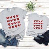 Red Hearts Metallic YOUTH Screen Print Transfer, two gray T-shirts featuring red metallic heart designs, casual denim jeans, and a small green potted plant on a white wooden surface.