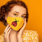 Lady Bug MASK/CAN HUGGER/POCKET Screen Print Heat Transfer, a yellow fabric face mask with a ladybug design on a polka dot background worn by a woman with red hair and blue eyes against an orange background.