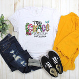 Your Grace Sublimation Transfer T-shirt with colorful floral and birds design and inspirational Bible verse, displayed with denim jeans, yellow long sleeve shirt, and black Vans sneakers on wooden background.