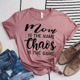 Mom Is The Name Chaos Is The Game Screen Print Heat Transfer T-Shirt, pink casual tee with black text and knot at the bottom, surrounded by jeans and brown shoes on a striped background.