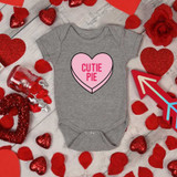 Cutie Pie Candy Heart INFANT Screen Print Heat Transfer on a gray baby onesie surrounded by red decorative hearts and Valentine's Day-themed items.