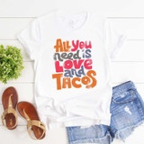 All You Need Is Love And Tacos Sublimation Transfer T-shirt with colorful text design, laid flat with denim shorts, sandals, and a small green plant on white wooden background.