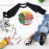 100 Days Sharper Sublimation Transfer, a graphic T-shirt with a cactus and sunset design, laid out with Crayola crayons, scissors, and casual denim and sneakers on a white wooden surface.