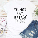 I'm Not Fat Sublimation Transfer, white t-shirt with black text saying 'I'm Not Fat I'm Easy To See', displayed with denim shorts and white sneakers on a wooden background.