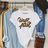 Walk By Faith Sublimation Transfer, white T-shirt with black and gold design hanging on a clothing rack.