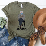 Bernie Mood FINAL STOCK Screen Print Transfer T-shirt, green casual shirt with an illustration of Bernie Sanders wearing a mask, sitting with crossed legs, holding mittens, underneath the text 'MOOD'.