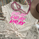 Stacked Pray Pink Screen Print Transfer, a beige t-shirt with pink lettering and prayer phrase print, dressed with pink beaded necklaces, placed on a multicolored woven rug with white crochet decor and a beige hat.