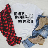 Home Is Where We Park It Sublimation Transfer, white T-shirt with a camper trailer graphic and text, paired with ripped blue jeans, a beige ankle boot, and a red plaid shirt, arranged on a beige textured surface.