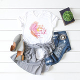 I Can Do All Things Through Christ Rose Sublimation Transfer, T-shirt with inspirational Bible verse and pink rose graphic, paired with striped shirt, distressed jeans, grey flats, black book with green plant on top, on white wooden background.