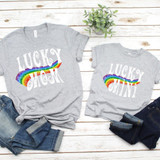 Lucky Mini Rainbow Screen Print Heat Transfer t-shirts, grey casual shirts with rainbow design and white 'LUCKY' text, laid on wooden surface with jeans and a small green plant.