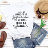 Jesus Take The Wheel Sublimation Transfer, white T-shirt with black and cursive text, laid flat on a white wooden surface with accessories including a straw hat, sunglasses, pink tulips, and casual sandals.