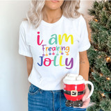 I Am Freaking Jolly Screen Print, a woman wearing a white T-shirt with colorful text and Christmas ornaments graphic, holding a Santa Claus themed mug filled with marshmallows, next to a decorated Christmas tree.
