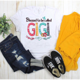Blessed To Be Called Gigi Sublimation Transfer, a colorful graphic t-shirt with floral and decorative accents laid flat alongside a pair of black Vans sneakers, a yellow jacket, distressed jeans, and a small green plant on a light wooden surface.