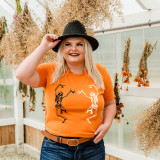 Dancing Skeletons Screen Print Heat Transfer, a woman wearing an orange T-shirt with dancing skeleton print, in a greenhouse with dried flowers hanging around.