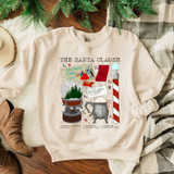 The Santa Clause DTF Transfer, graphic Christmas-themed design on a white sweatshirt featuring Santa's setup including a mystery date game, North Pole sign, snowglobe, business card, and hot cocoa mug.