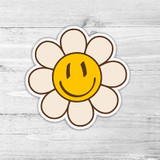 Smiley Flower Retro Die Cut Sticker, cheerful flower with a smiley face at the center and cream-colored petals on a wooden background.