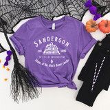 Sanderson Witch Museum WHITE DTF Transfer, showcasing a purple t-shirt with white print of the Sanderson Witch Museum logo, surrounded by Halloween-themed decorations like pumpkins, purple bows, and a black web-patterned cloth.