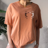 Dancing Skeletons HAT/POCKET Screen Print Heat Transfer, a t-shirt featuring a design of two dancing skeletons with raised arms and posed legs, in black and white, on an orange t-shirt.