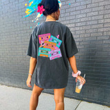 90s Cassette Tapes Screen Print Heat Transfer, colorful cassette tapes with nostalgic text and graphics on a black T-shirt worn by a woman standing against a brick wall.
