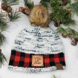 Winter Is Stupid TAN Leather Patch, cozy knit beanie with faux fur pom-pom, surrounded by pine branches and pinecones in a winter setting.