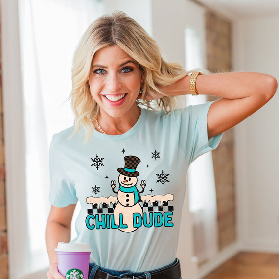 Chill Dude Snowman DTF Heat Transfer, a woman in a light blue t-shirt featuring a snowman graphic and the text 'Chill Dude' with snowflakes, holding a coffee cup and smiling.