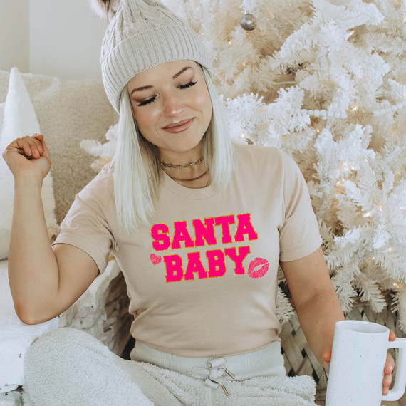 Santa Baby Faux Chenille Screen Print Heat Transfer, woman wearing a cream t-shirt with 'Santa Baby' pink and gold text and lips graphic, sitting near a decorated white Christmas tree.