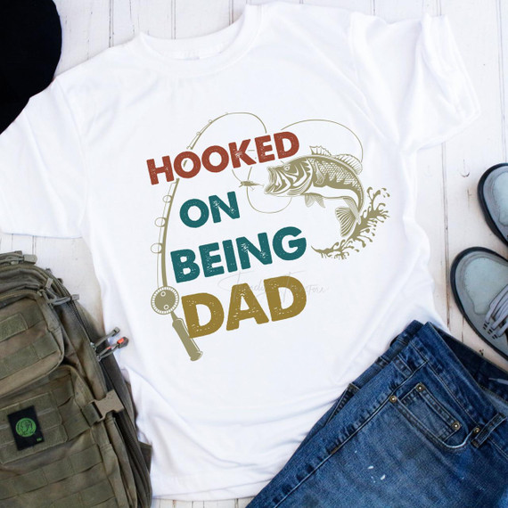 Hooked On Being Dad Fishing Sublimation Transfer, a digital design on a white T-shirt featuring a fishing theme with a fish, fishing line, and colorful text.