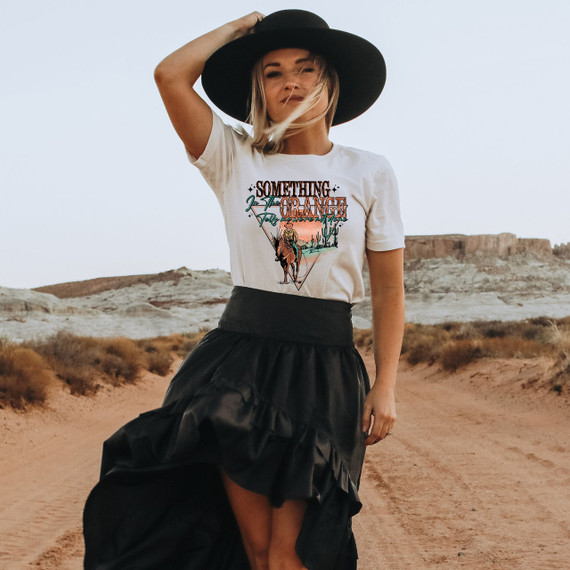 Something In The Orange Screen Print Heat Transfer, a woman wearing a white t-shirt with the design, standing in a desert landscape with rocky formations and clear sky.
