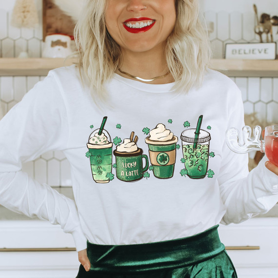 Lucky Lattes DTF Heat Transfer, image of a woman wearing a white sweatshirt with illustrations of four different green-colored latte drinks with shamrocks, representing St. Patrick's Day or Irish theme.