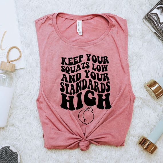 Keep Your Squats Low And Your Standards High DTF Heat Transfer, pink t-shirt with motivational slogan and a peach graphic, laid flat on a white fluffy surface with surrounding fashion and beauty accessories.