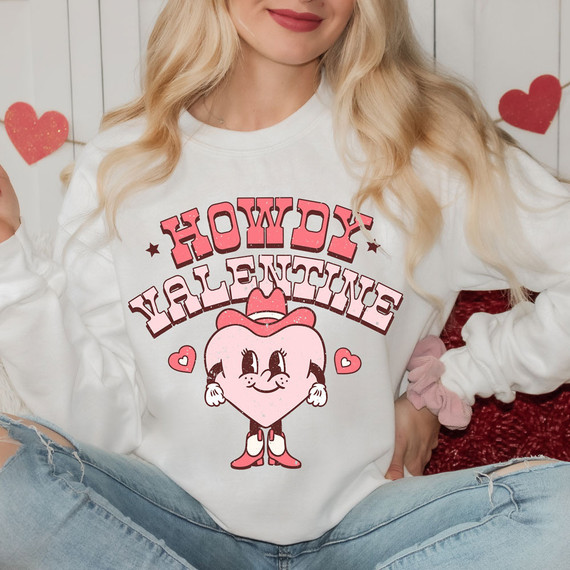 Howdy Valentine Pink DTF Heat Transfer, pink heart-shaped character with cowboy hat and boots, surrounded by pink and red hearts on a white sweatshirt.