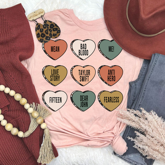Taylor Candy Hearts DTF Heat Transfer, colorful heart-shaped designs with words like 'MEAN,' 'BAD BLOOD,' 'LOVE STORY,' and 'FEARLESS' on a pink t-shirt, surrounded by fashion accessories.