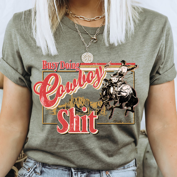 Busy Doing Cowboy Shit DTF Heat Transfer, humorous iron-on graphic design featuring a cowboy riding a bucking horse with a desert landscape background.