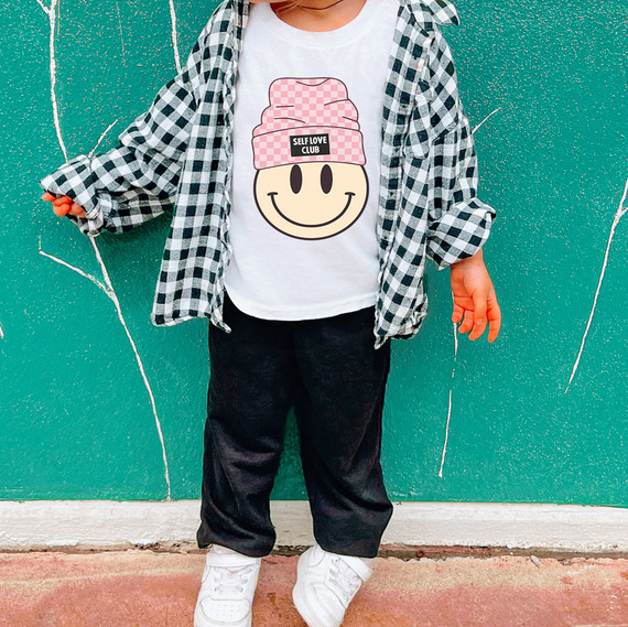 Self Love Club Beanie Sublimation Transfer, close-up of a child wearing a white t-shirt with a smiley face logo wearing a pink checked beanie, and a black and white checkered shirt, standing against a turquoise wall with white graffiti.