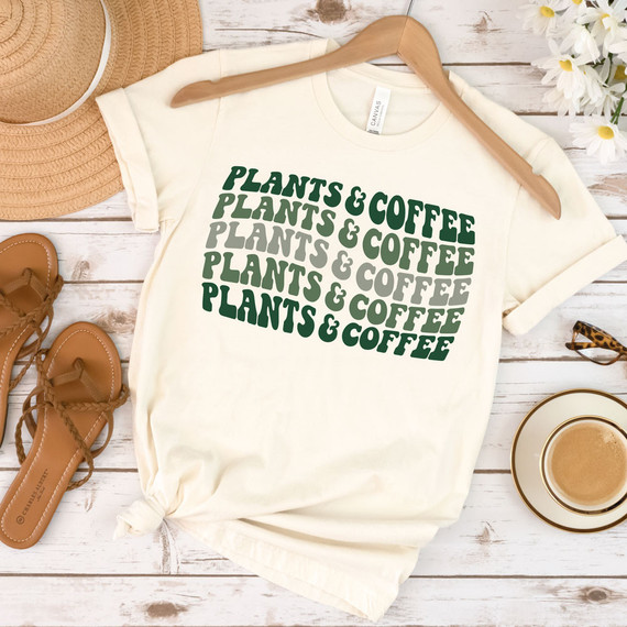 Plants & Coffee DTF Heat Transfer, a graphic t-shirt with layered green and gray text design, on white fabric, surrounded by accessories on a rustic white wooden surface.