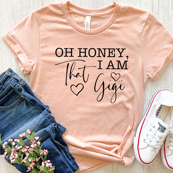 Oh Honey, I Am That Gigi DTF Heat Transfer, pink T-shirt with black text and hearts design, laid flat with denim jeans, pink flowers, and white sneakers.