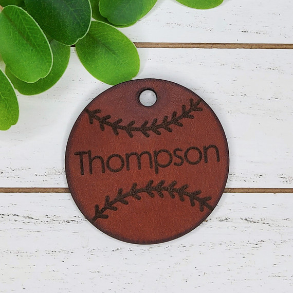 Personalized Leather Baseball Softball Keychain, brown leather keychain with black embroidered baseball stitch design and customizable name, with a small hole for keyring attachment, on white wooden surface with green leaves in the background.