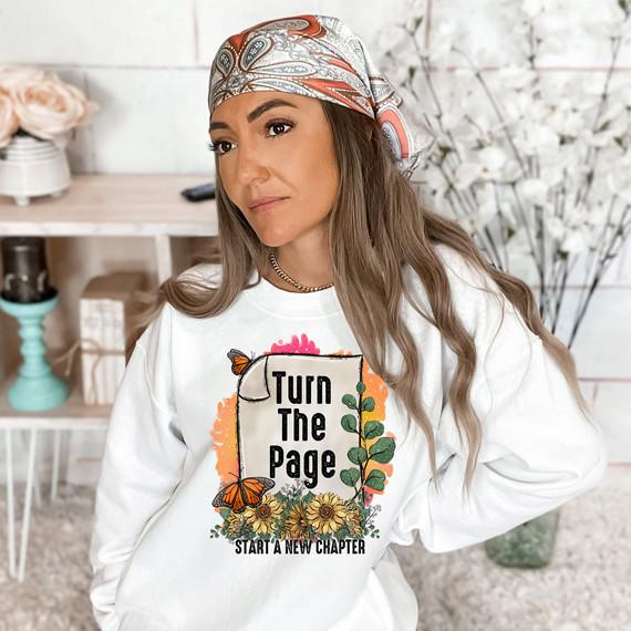 Turn The Page Start A New Chapter DTF Heat Transfer, a woman wearing a white sweatshirt with the design, floral and butterfly accents, and a colorful headscarf.