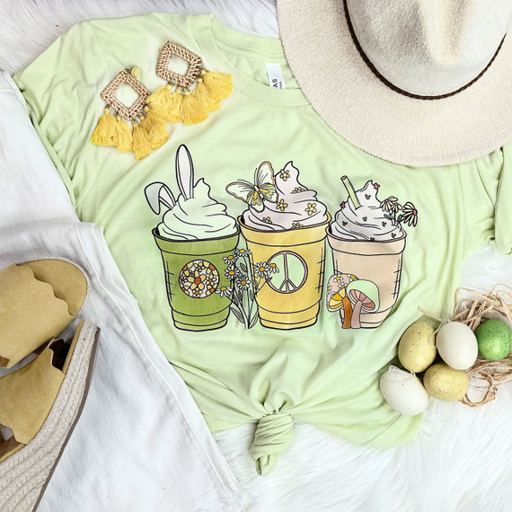 Easter Latte Screen Print Heat Transfer, colorful illustration of three whipped cream beverages with bunny ears, butterflies, flowers, and mushrooms on a pastel green t-shirt