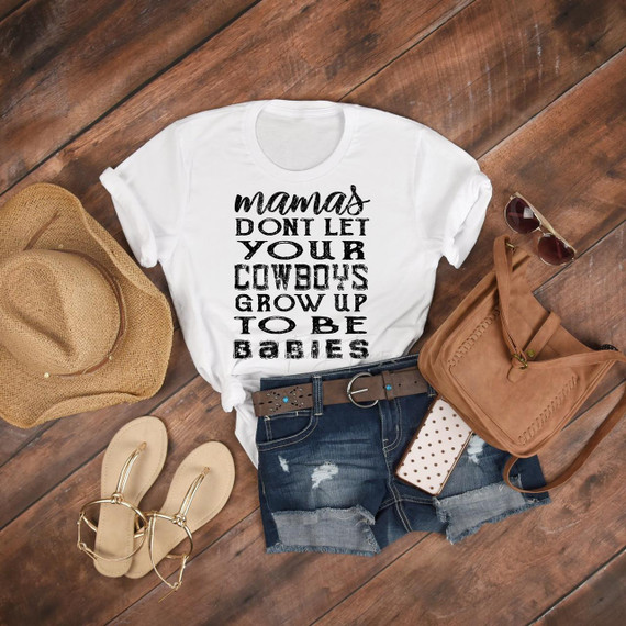 Mamas Don't Let Your Cowboys Grow Up To Be Babies Sublimation Transfer, product image of a white T-shirt with black text, paired with denim shorts, a straw hat, beige sandals, sunglasses, a brown handbag, and a polka-dotted phone case on a wooden floor.