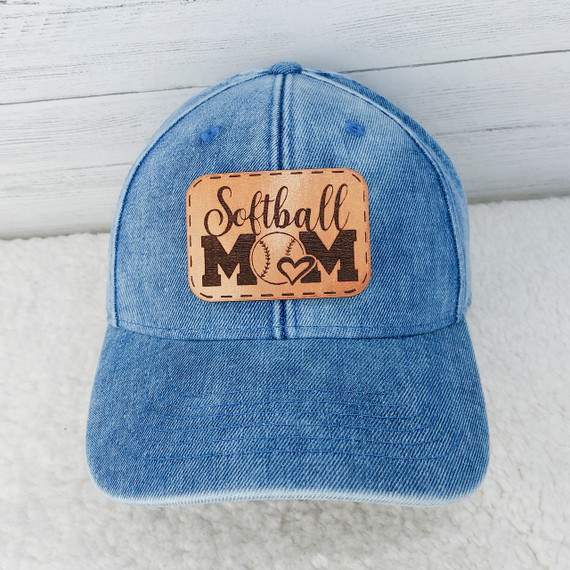 Softball Mom Leather Hat Patch, blue denim baseball cap with a tan leather patch embroidered with the words 'Softball Mom' and a baseball graphic surrounded by a stitched border.