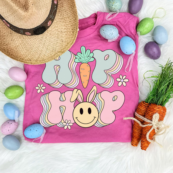 Hip Hop Retro Easter DTF Heat Transfer, colorful Easter-themed design on pink t-shirt with bunny, carrot, and flowers surrounded by pastel-colored Easter eggs and a straw hat with floral band, on white faux fur background.
